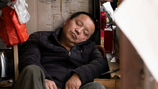 A man sitting down having a nap on the side of a busy street.