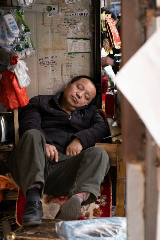 A man sitting down having a nap on the side of a busy street.