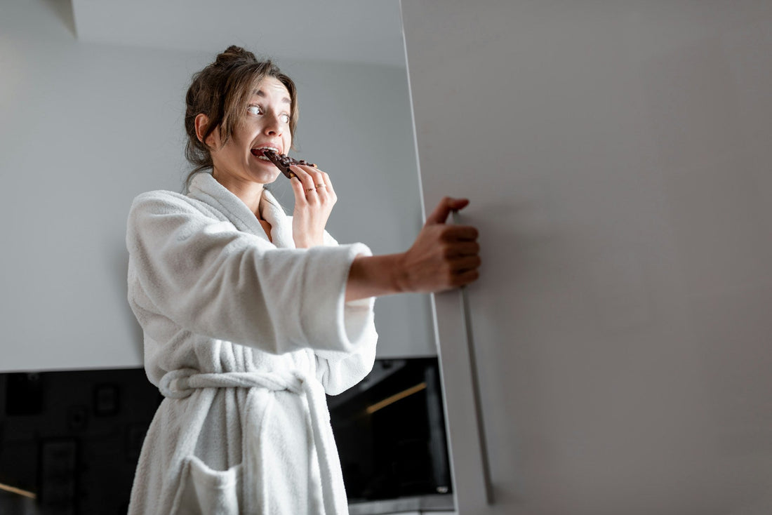 A woman in a dressing gown eating chocolate from a fridge
