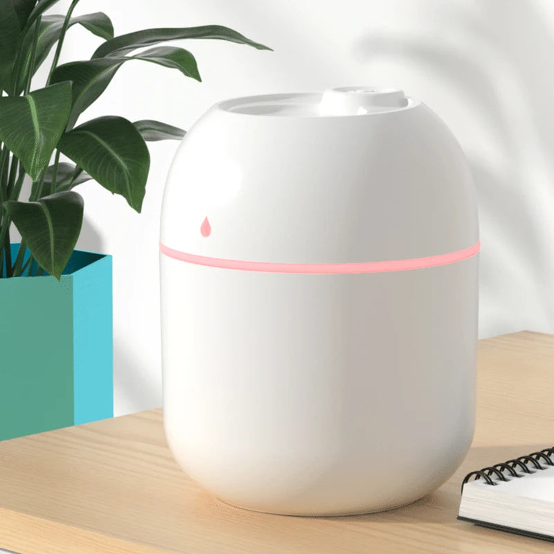 White humidifier with a pink light on a wooden surface next to a plant and notebook.
